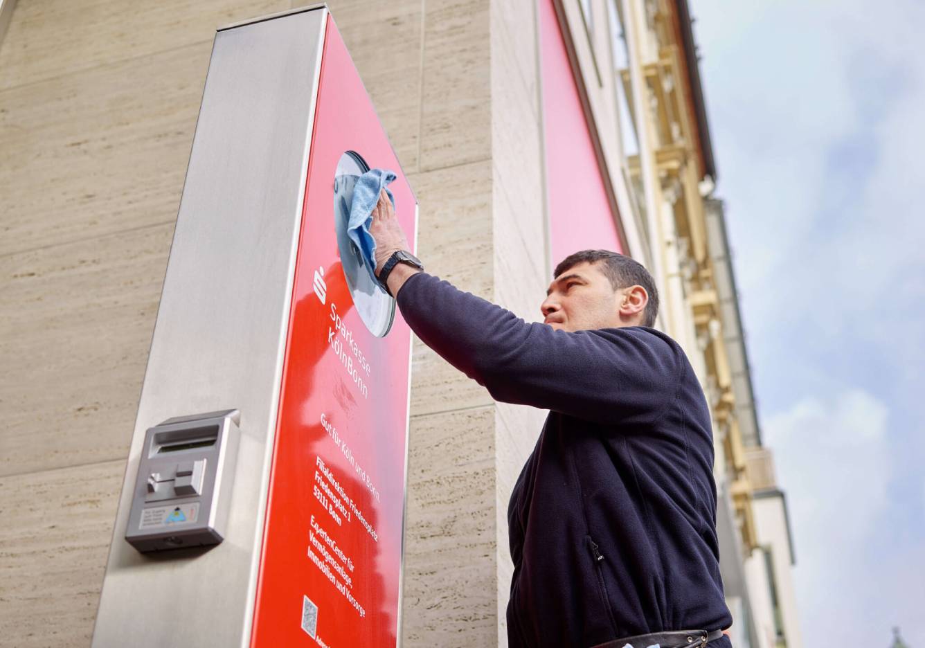 Gebäudereiniger von Clean Excellence reinigt das Schild am Eingangsbereich einer Sparkassenfilliale.